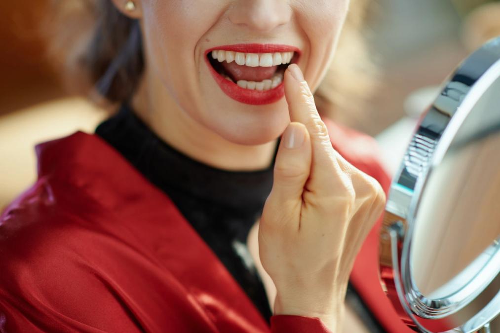 Smiling woman in red touching her teeth while looking in a mirror.