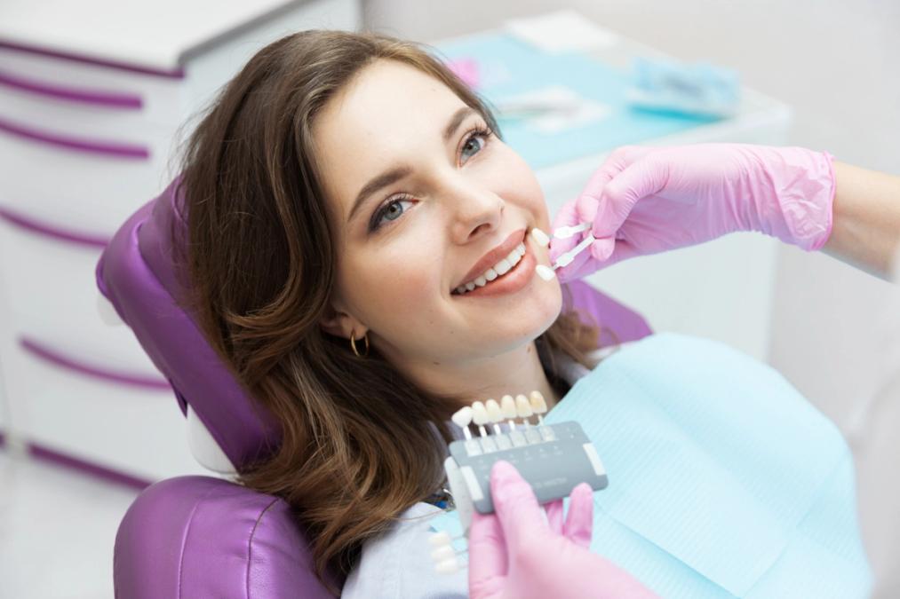 Smiling woman in dental chair having tooth shade matched by a gloved hand.