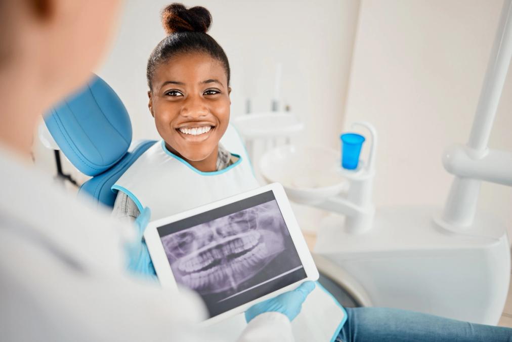 A smiling woman in a dental chair looks at a tablet displaying her dental X-rays, held by a dentist in the foreground.
