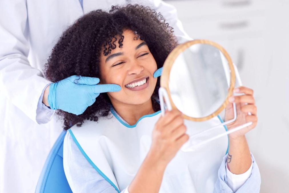 A happy woman in a dental chair looks at her smile in a mirror while a gloved hand points to her teeth.