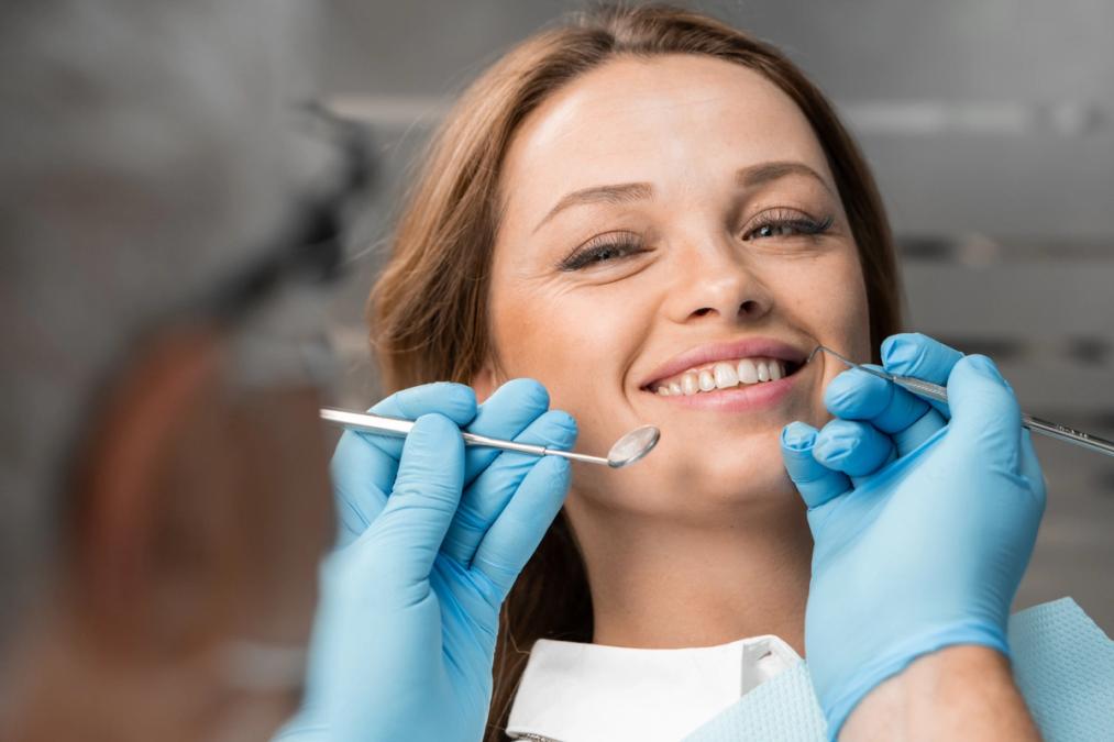 A smiling woman at a dental check-up, with a dentist's gloved hands holding tools near her mouth.