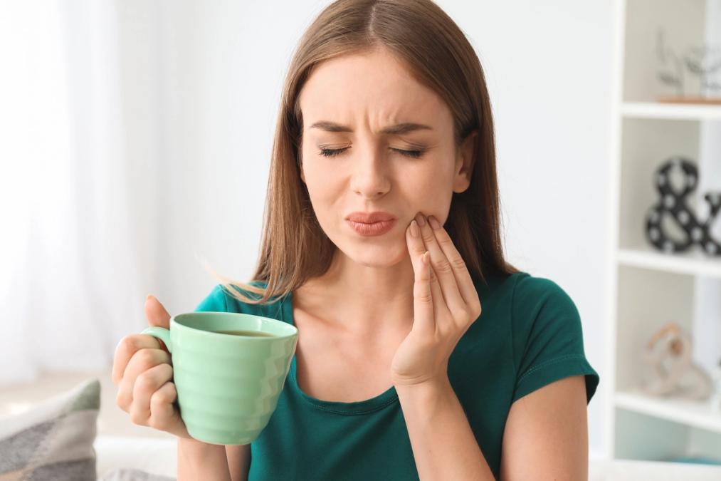 A woman winces in pain, holding her cheek while holding a green mug.
