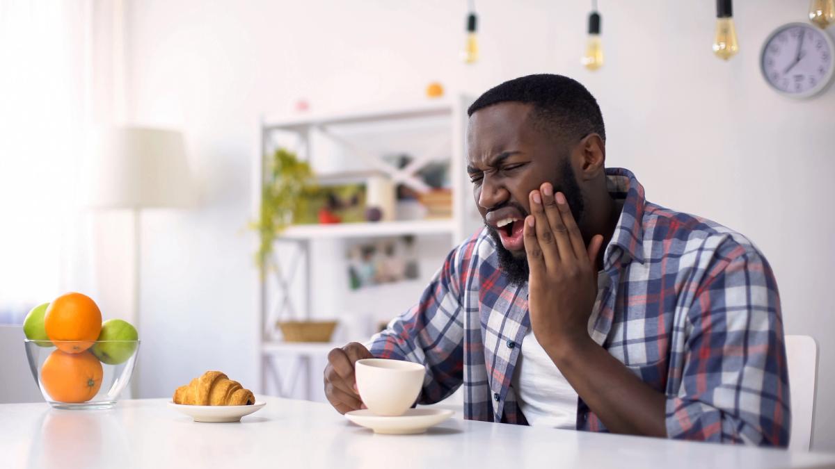 A man grimaces in pain, holding his cheek as if from a toothache, while sitting at a table with a cup of coffee.