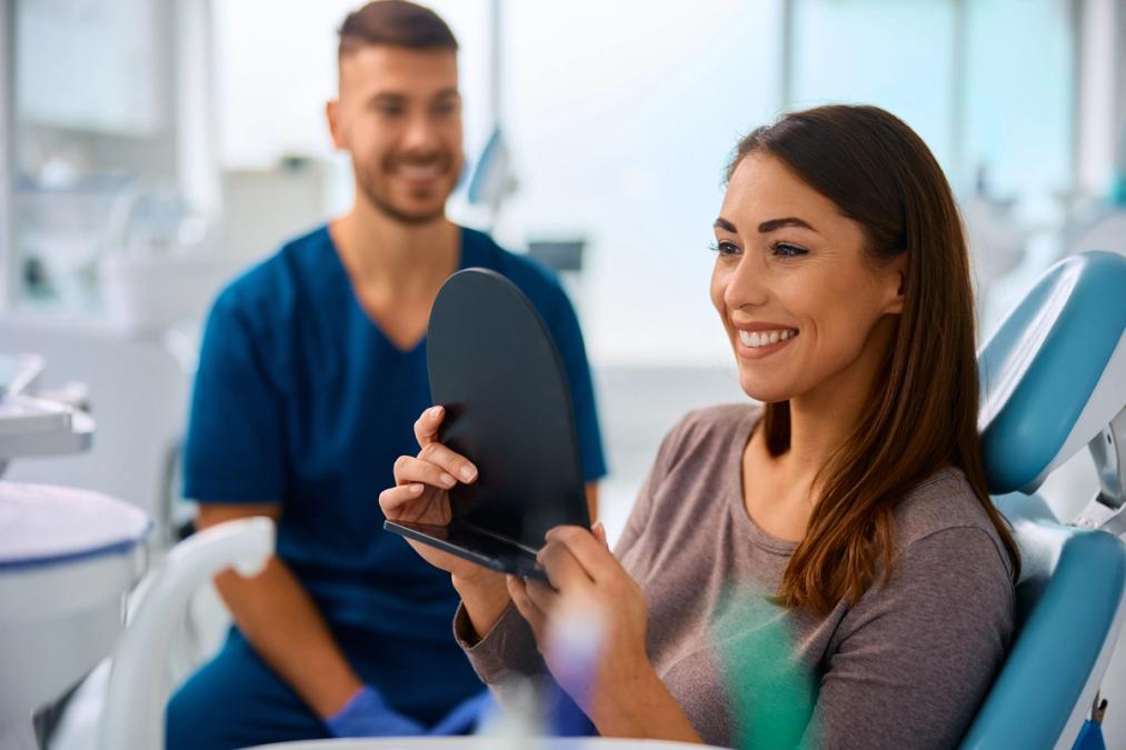 Smiling woman in a dental chair looks at her reflection in a mirror, with a smiling dentist in the background.