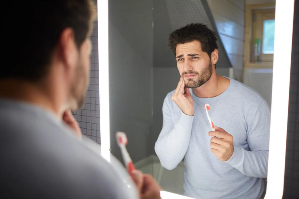 Man in a mirror holding a toothbrush, pressing his hand to his cheek with a pained expression.