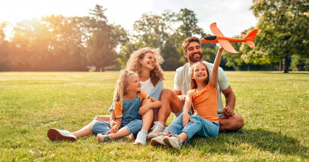 A happy family of four sits on grass in a sunny park, with a child holding an orange toy airplane.